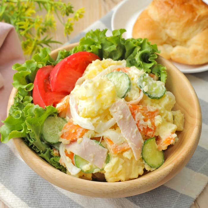 サラダボウルに入ったポテトサラダ、レタス、トマト、キュウリ、ハムが盛り付けられた料理。横にクロワッサンがある。