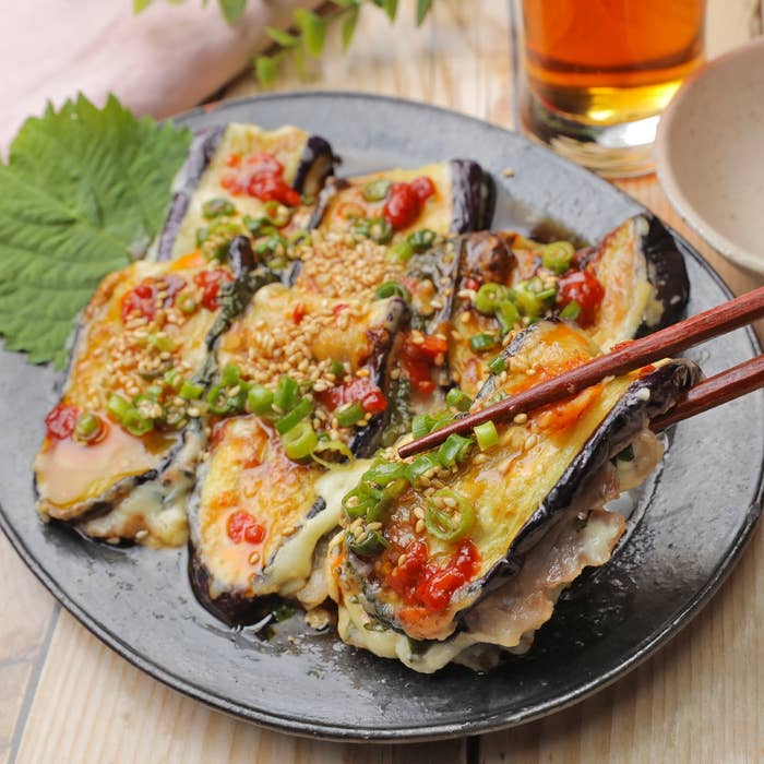焼き茄子に辛いソースとゴマ、ネギが載せられた料理が盛り付けられた黒い皿。隣に茶色の飲み物のグラス。
