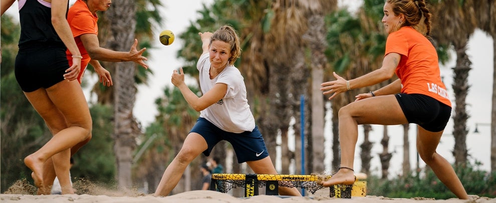 People playing a beach game, energetically hitting a ball towards a small yellow and black net on the sand, with palm trees in the background