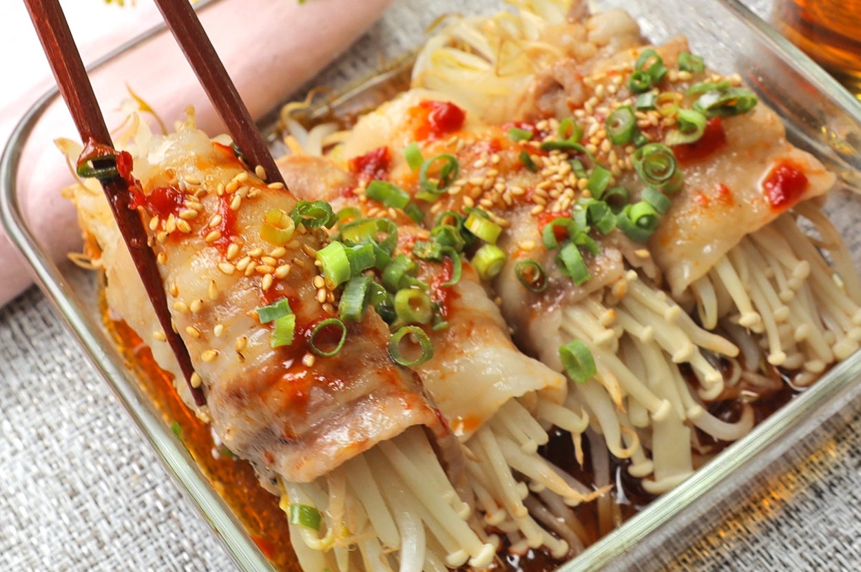 ガラス製の皿に盛られたエノキ茸の棒が豚肉で巻かれ、上にネギとごまが振りかけられた料理。箸が一つ持ち上げている。
