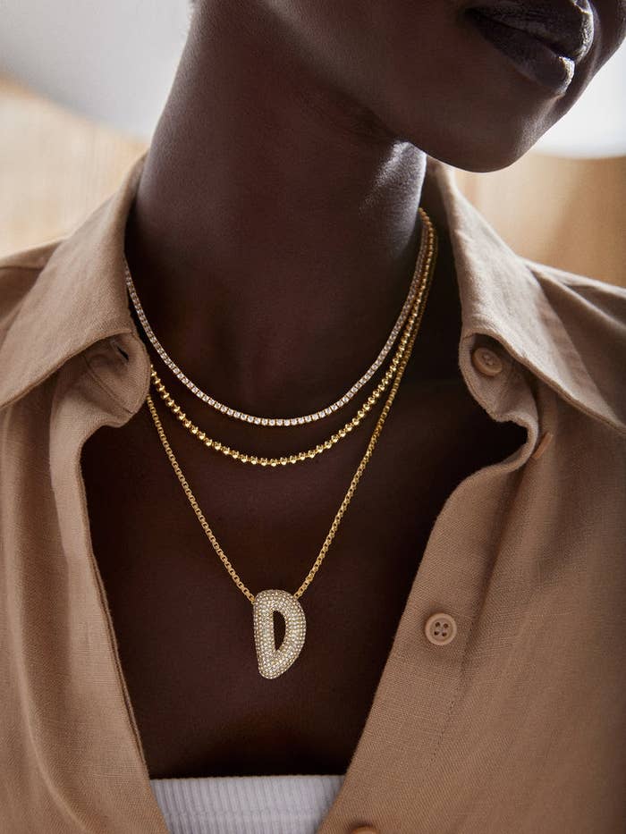 A idiosyncratic wears layered golden necklaces featuring a ample D pendant implicit a casual button-up shirt.