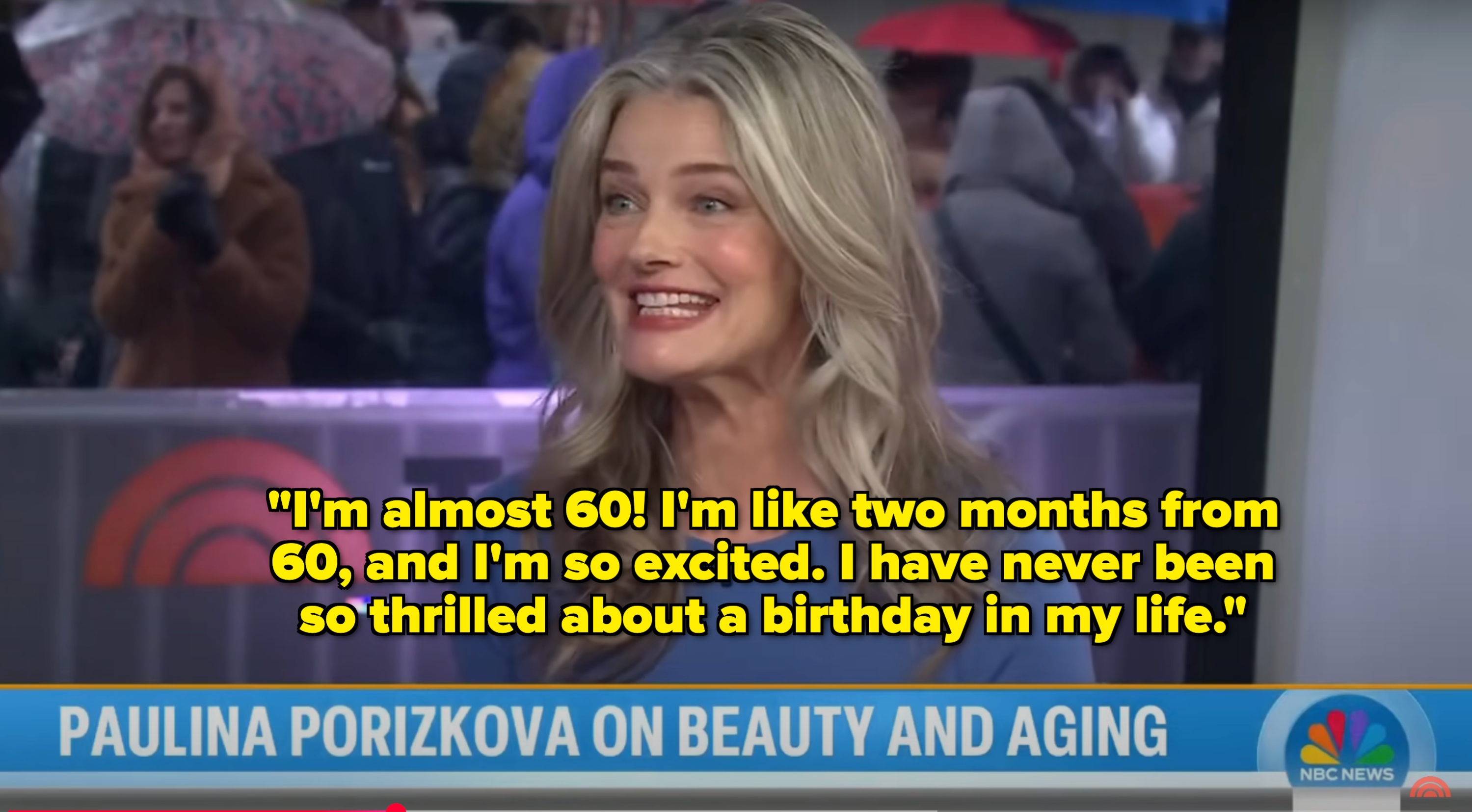 Kvinne som smiler under et TV -intervju om skjønnhet og aldring, med bakgrunn av uskarpe tilskuere og en showlogo delvis synlig