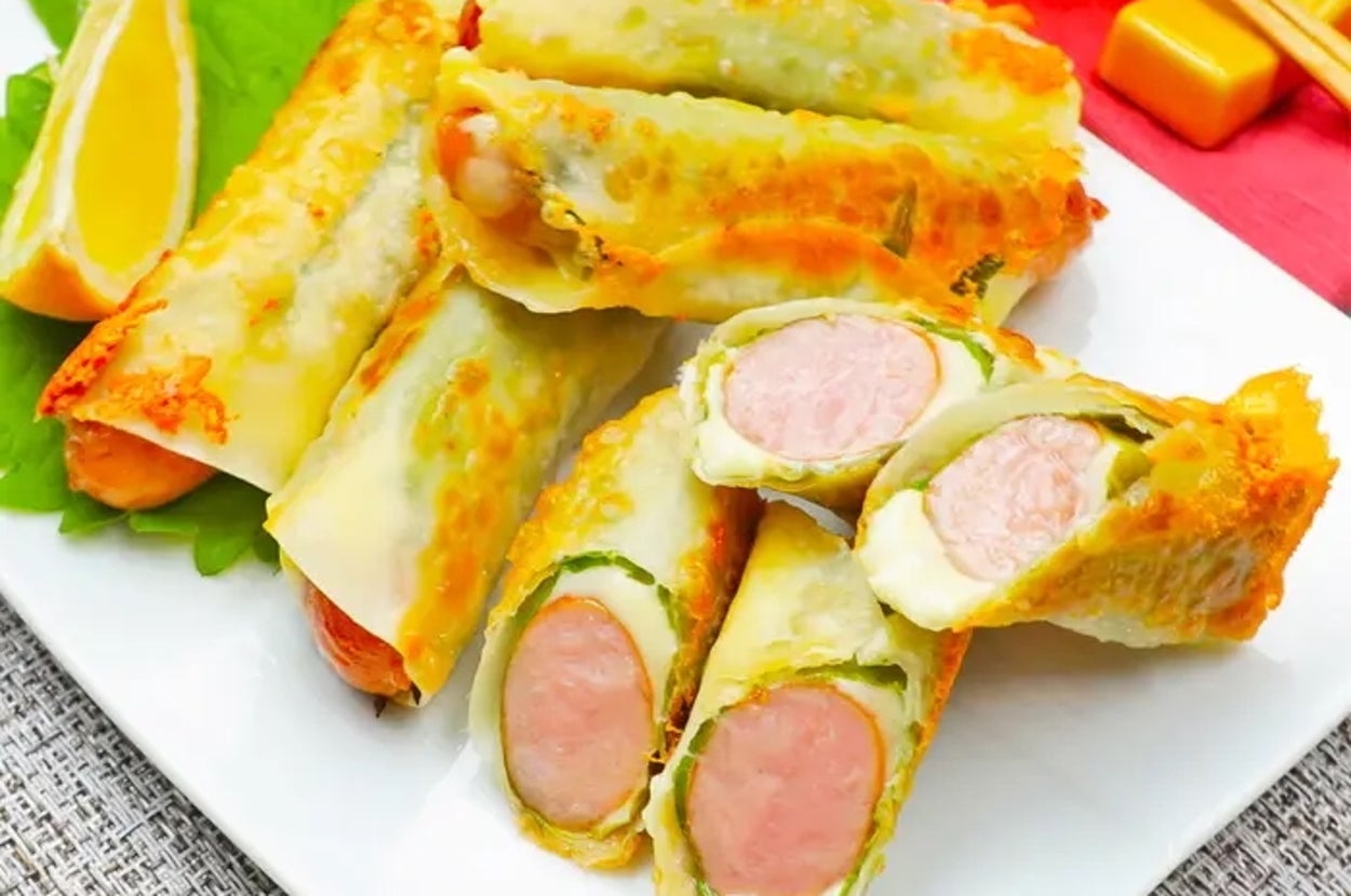 白い皿に盛り付けられたソーセージ入り春巻き、レモンのくし形と飲み物が添えられた広告用料理の画像。