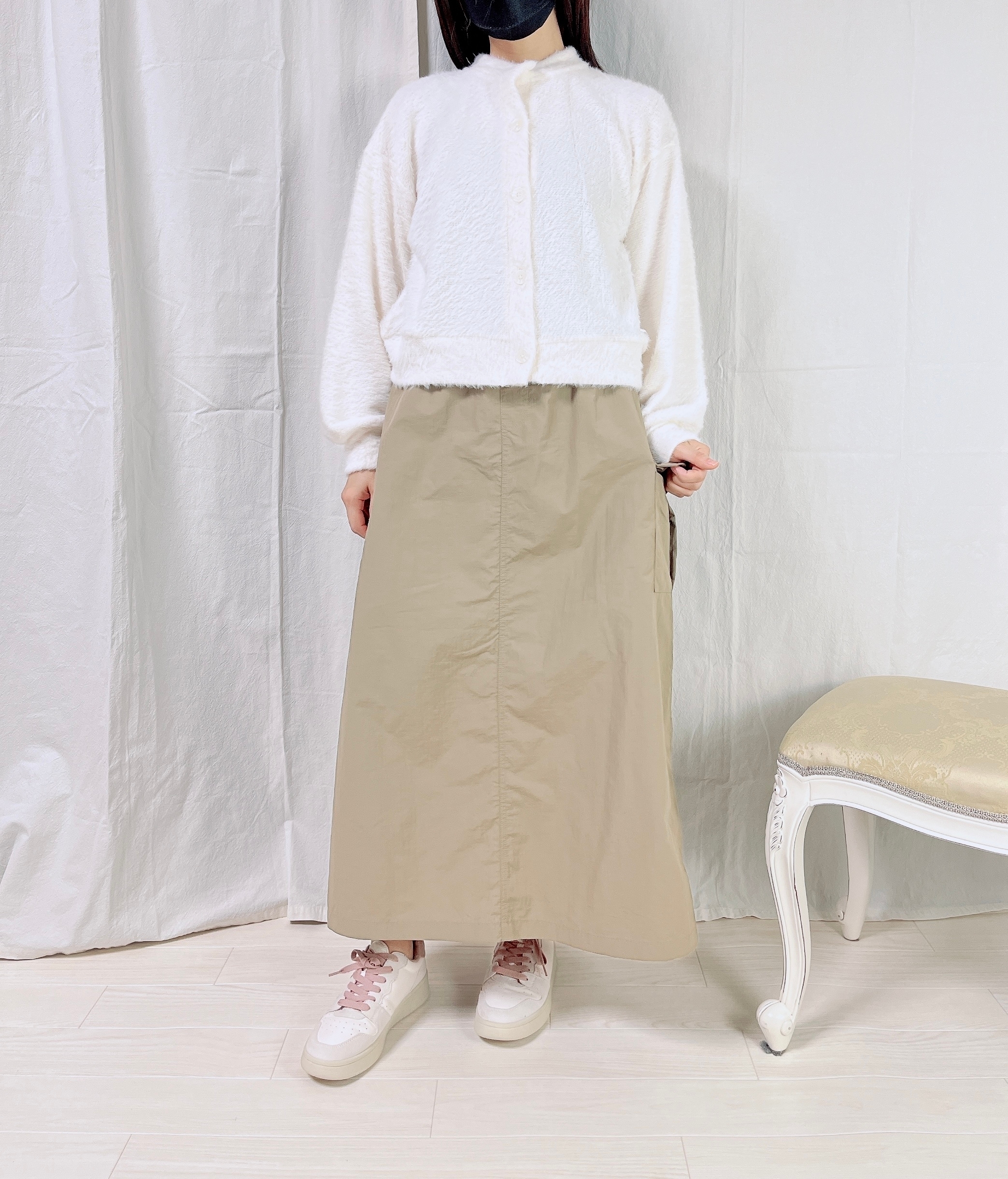 女性が白い長袖のトップスとベージュのロングスカートを着て立っている。顔は写っていない。横に椅子がある。
