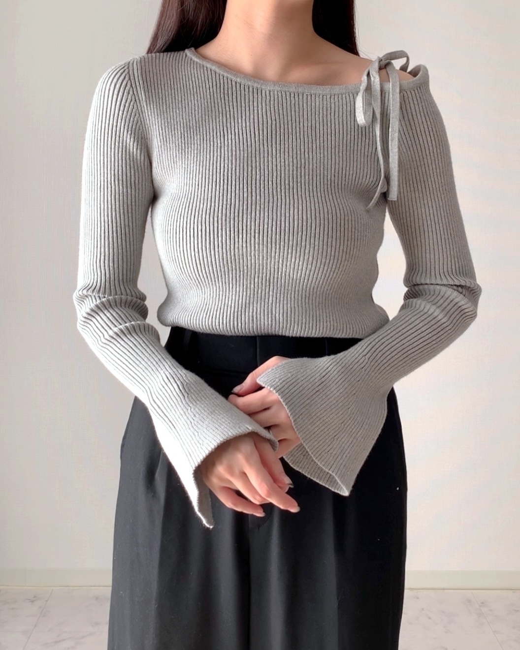女性が片袖にリボンのあるリブニットセーターと黒いボトムスを着ている。