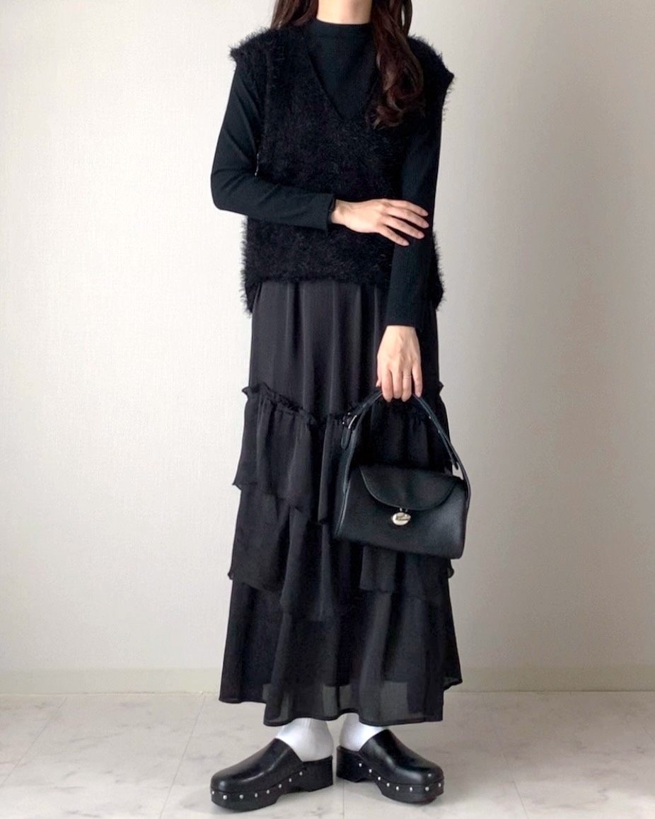 女性が黒のセーターとフリルスカートを着用し、黒のバッグを持っています。