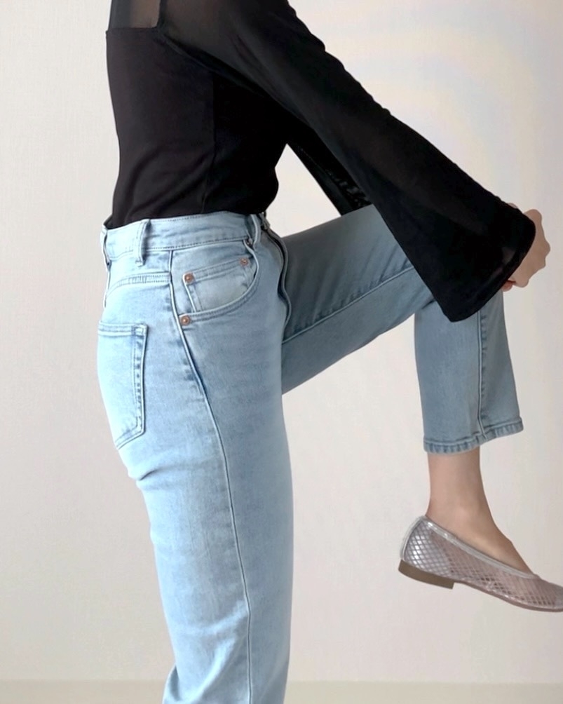 女性が片足を上げ、ライトウォッシュジーンズとベルスリーブの黒いトップスを着用している。
