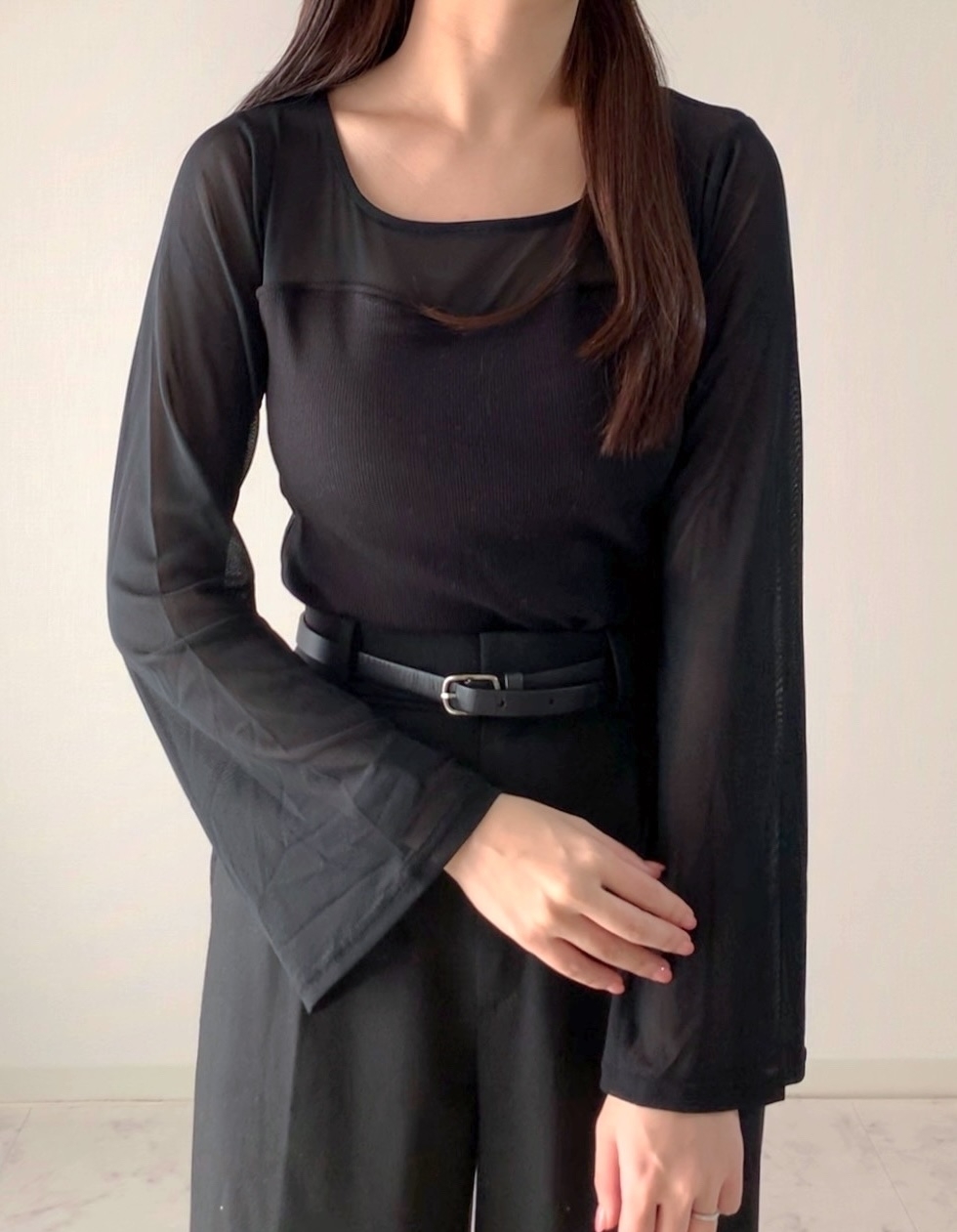 女性が長袖黒トップスとベルト付きパンツを着て立っている。