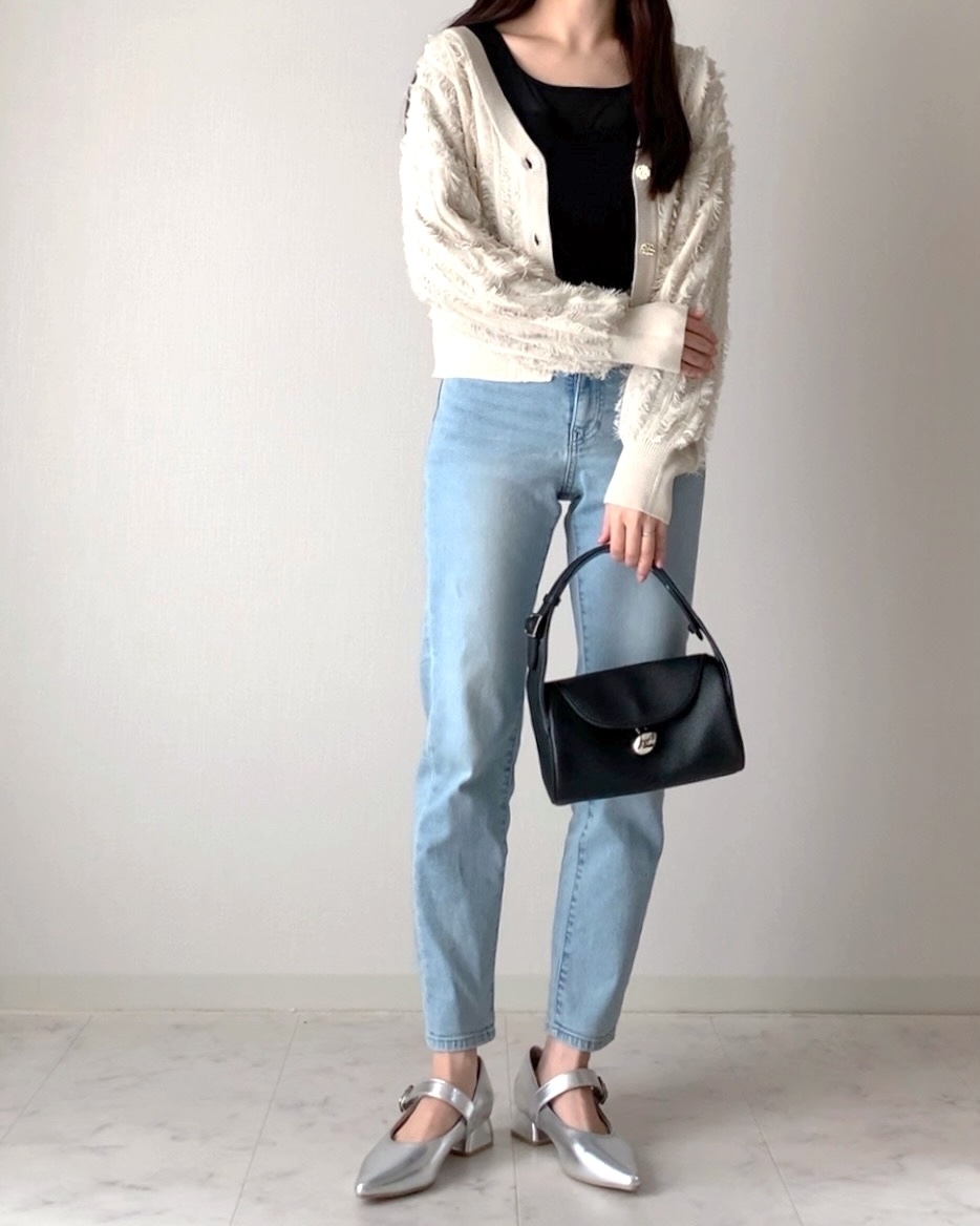 女性が黒のトップス、ベージュのカーディガン、デニムパンツ、シルバーの靴、黒のバッグを持って立っている。
