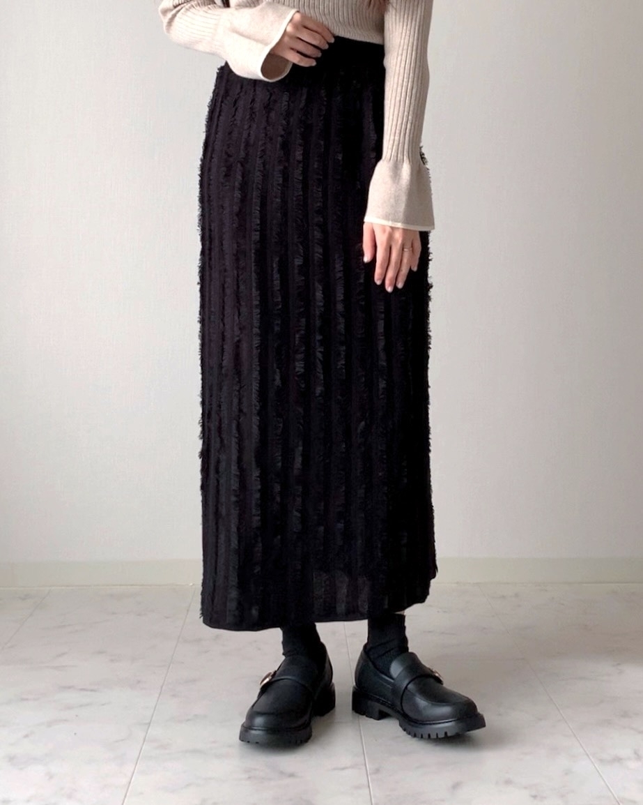 女性が、長袖のリブニットと、プリーツスカート、ローファーを履いて立っている。