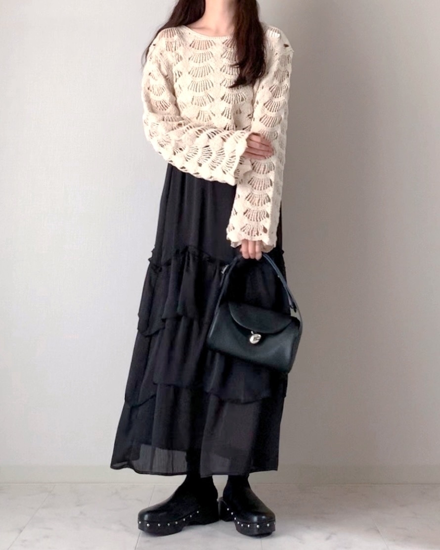 女性がニットトップとロングスカートを着て、バッグを持って立っている。
