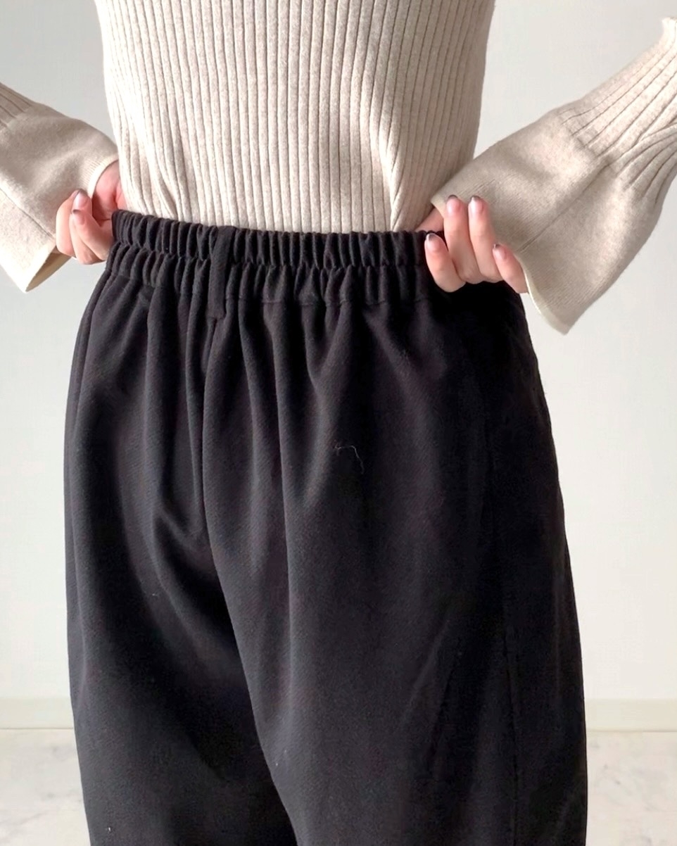 女性がゆったりとした黒いクロップドパンツのウエストをつかんでいる様子。リブニットのトップスを着用。
