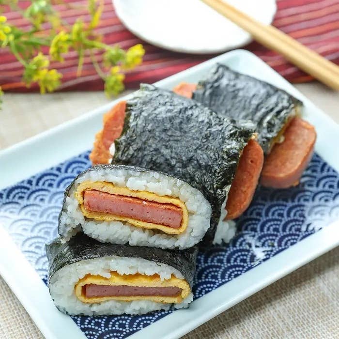 皿の上にのったスパムむすび。海苔で巻かれた米とスパム、薄焼き卵が挟まれている。背景に箸と花。