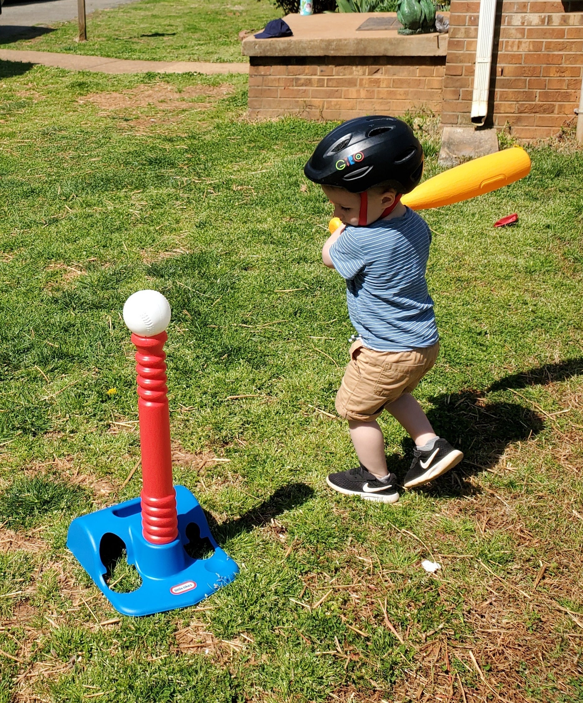 A young child in a helmet prepares to hit a ball with a plastic bat in a backyard setting