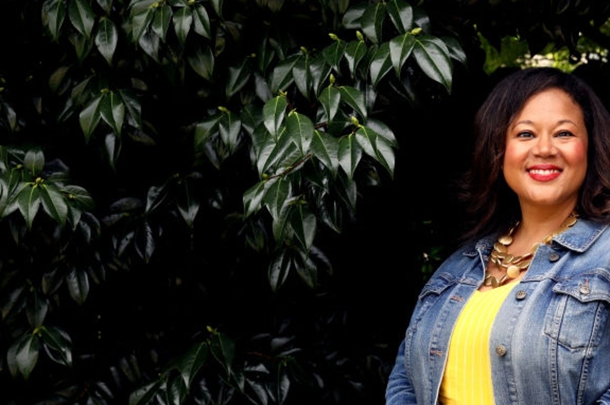 Person in a bright dress and denim jacket stands smiling, hands clasped, in front of lush green foliage