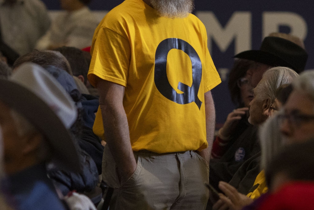 Person i en gul skjorte med en stor & quot; Q & quot; deltar på et overfylt innendørs arrangement, omgitt av sittende deltakere