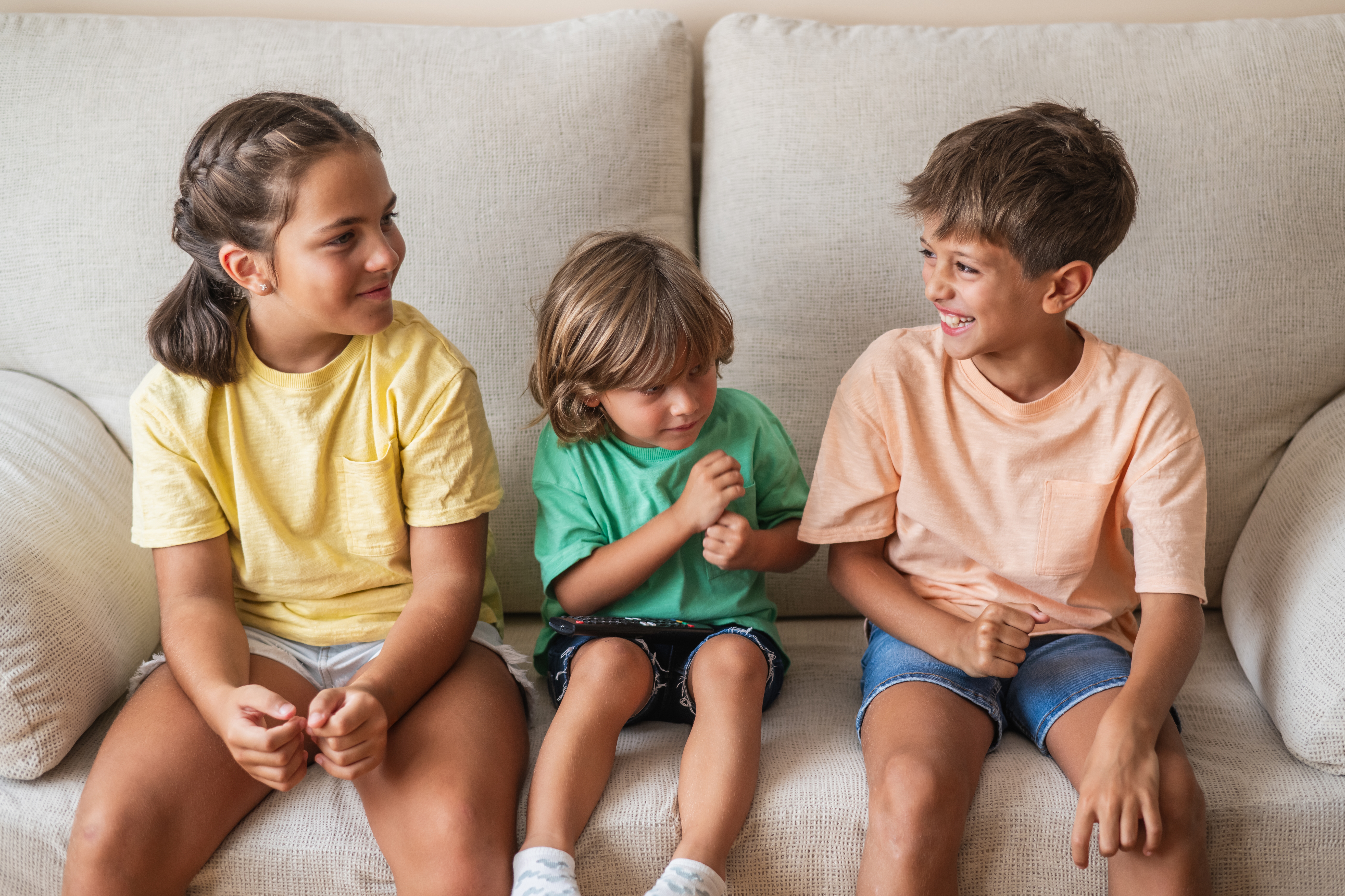 Tre barn som sitter på en sofa, smiler og chatter med hverandre, har på seg uformelle sommerklær