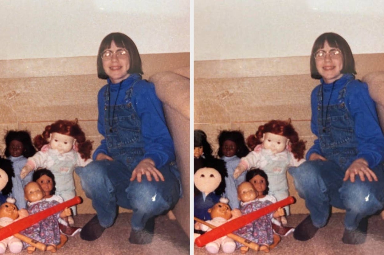 A person smiling while kneeling next to several dolls on a carpet, dressed in denim overalls and a long-sleeve top