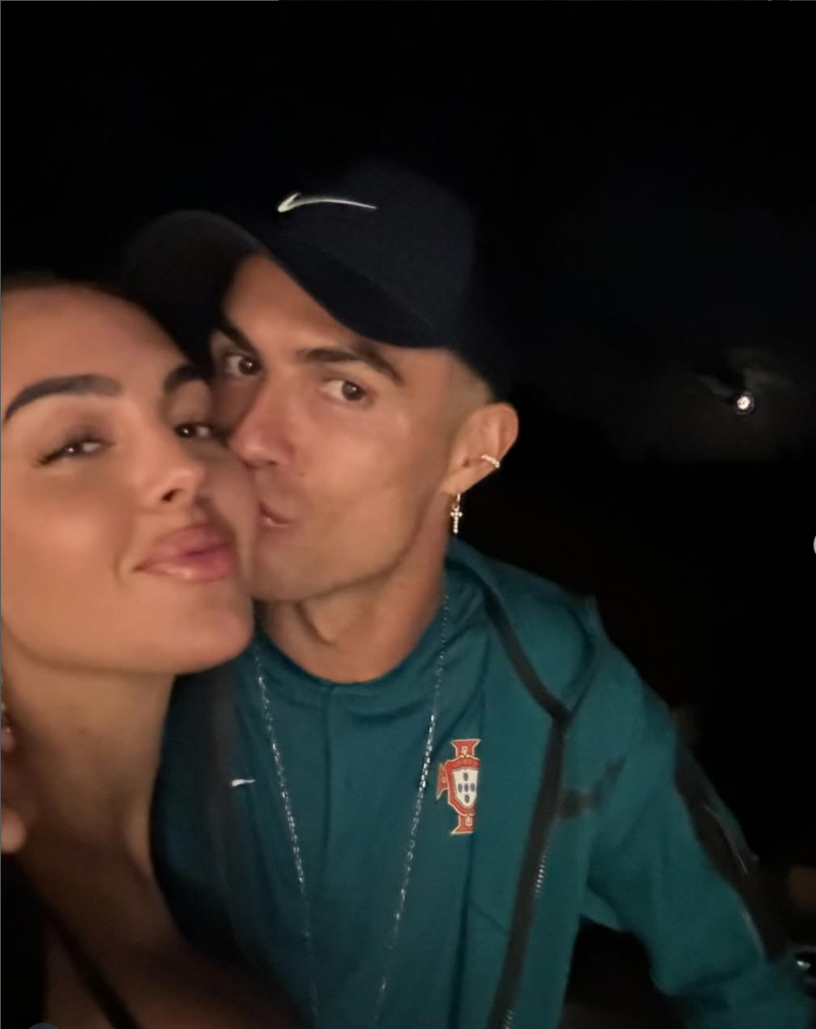 Two people taking a selfie; one wears sporty clothing, including a hat and earrings, and is kissing the other's cheek