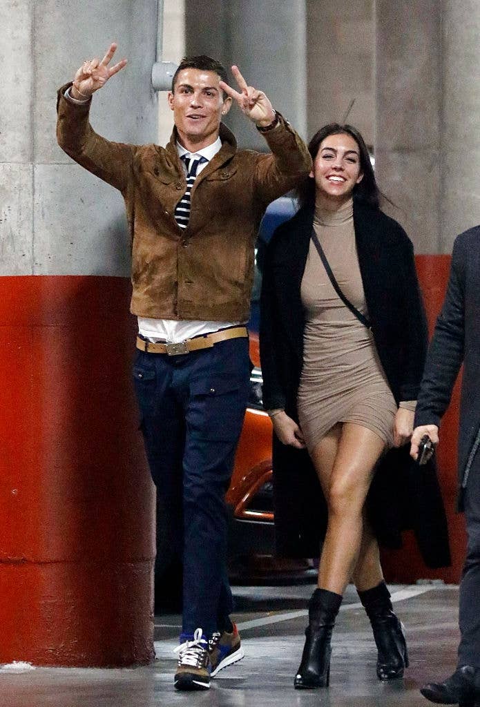 Two individuals smiling, one in a suede jacket and jeans, the other in a fitted dress and boots, walking with one raising their hands in a peace sign