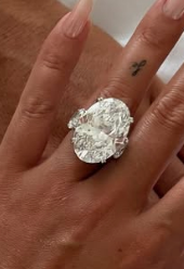 Two hands holding on a white bed, one with a large diamond ring and long nails. The image suggests intimacy or engagement