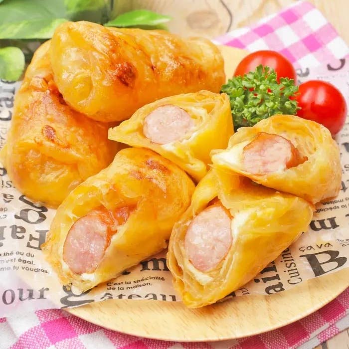 パイ生地にソーセージが巻かれた料理が皿に盛られている。パセリとトマトが添えられている。
