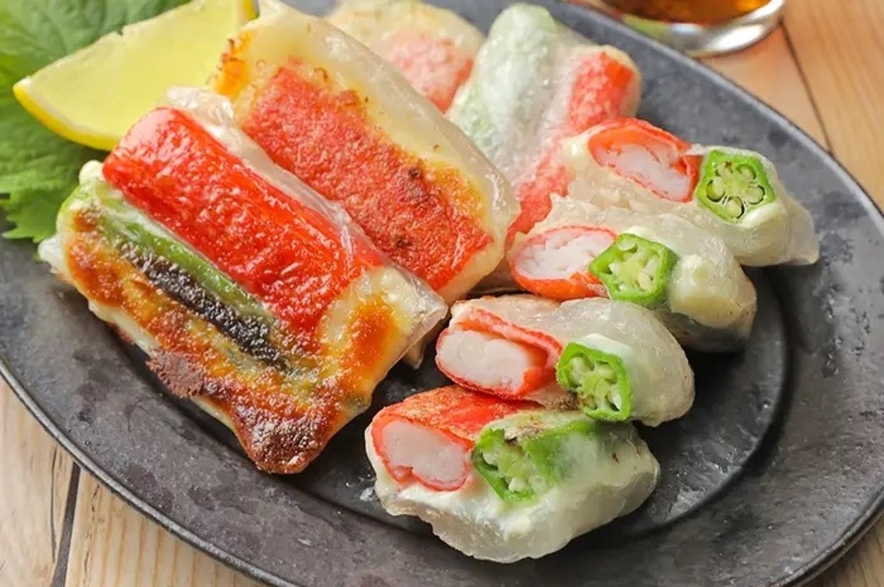 おいしそうな焼き餃子が黒い皿に盛られている。中にカニカマやオクラが入っており、レモン片と飲み物が添えられている。