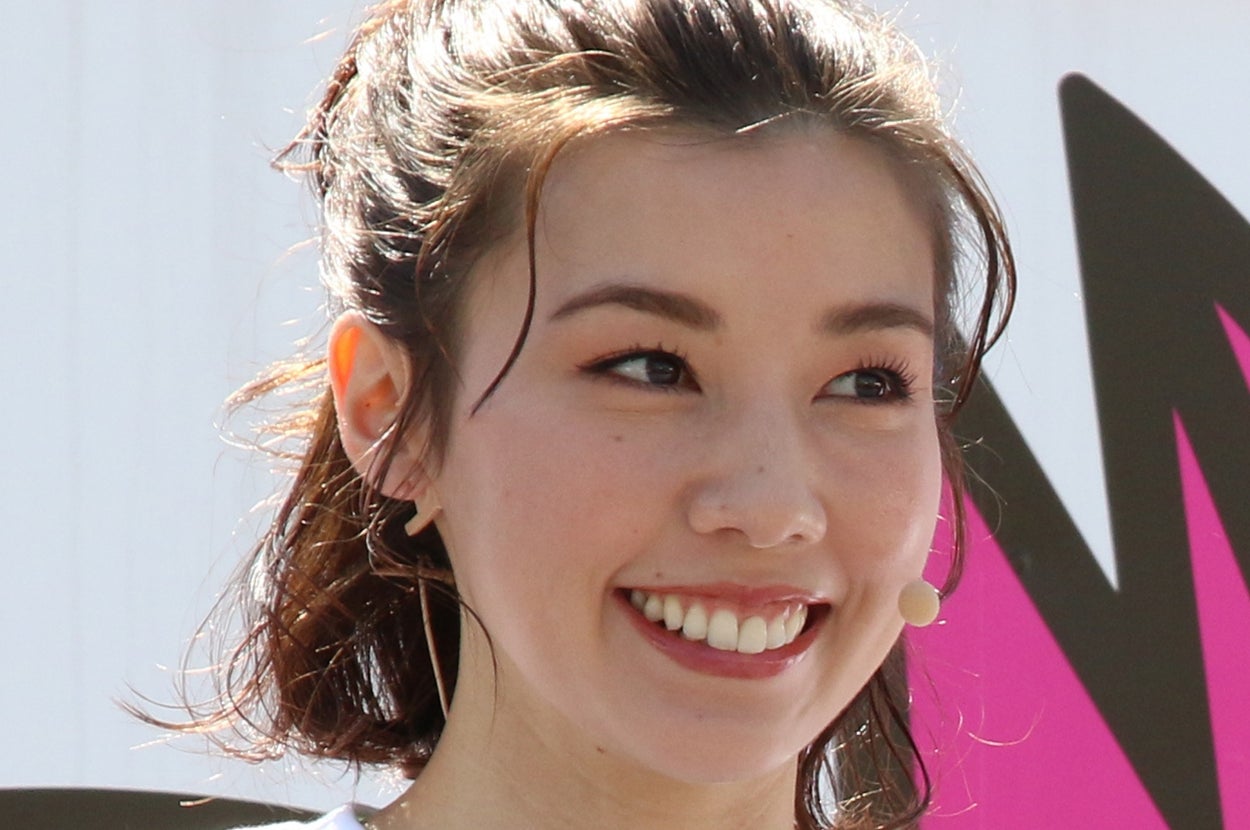 女性が白いTシャツを着て、笑顔でインタビューに答えている。髪は肩までの長さで、ピンで留めているようです。