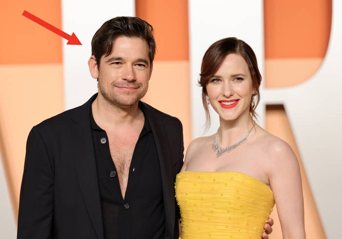 Jason Ralph in a blazer with an open shirt and Rachel Brosnahan in a strapless, textured dress and necklace, smiling on the red carpet