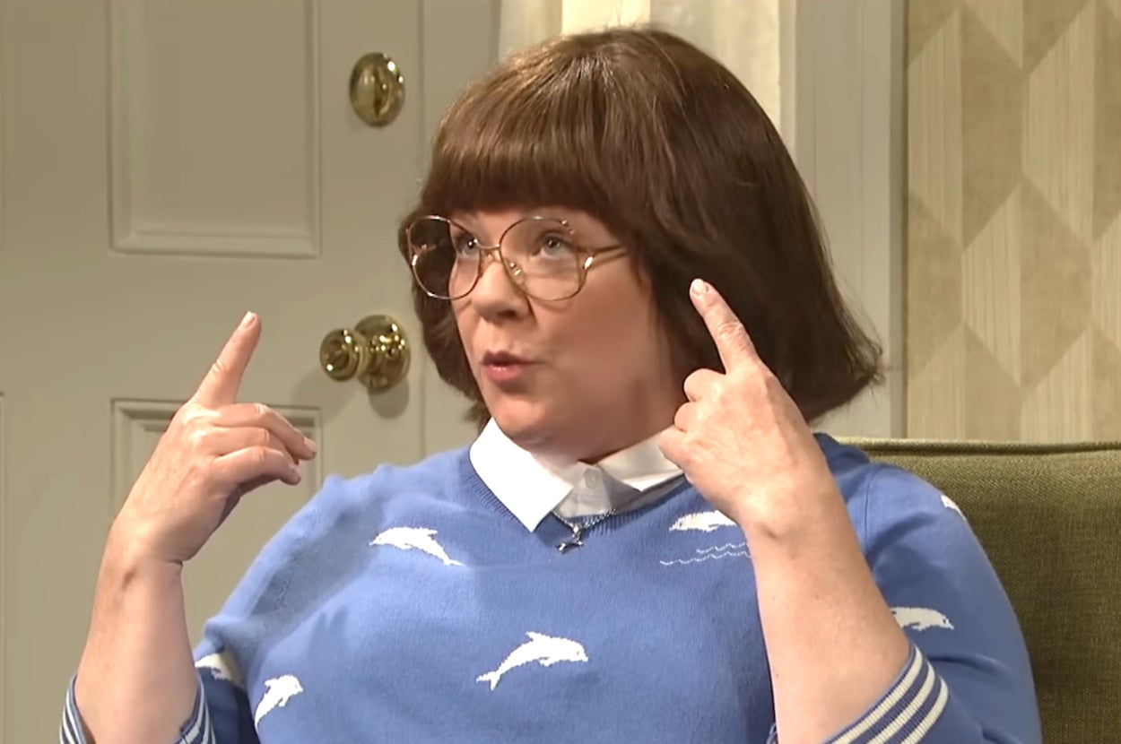 Person in retro-style glasses wears a sweater with dolphin patterns, gesturing emphatically with both hands pointing upward