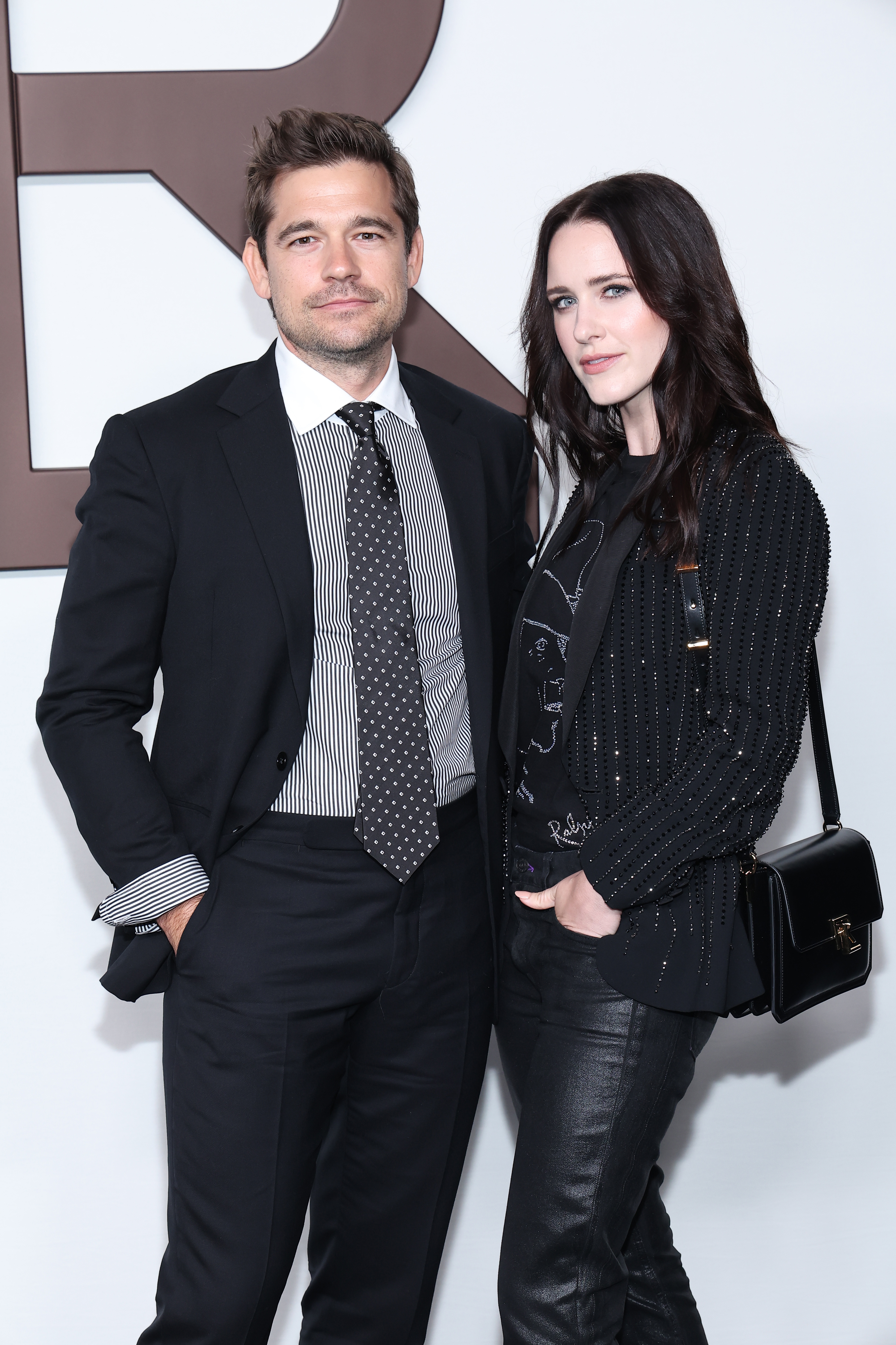 Jason Ralph in a pinstripe suit with a polka-dot tie and Rachel Brosnahan in a textured blazer over a graphic tee and leather pants