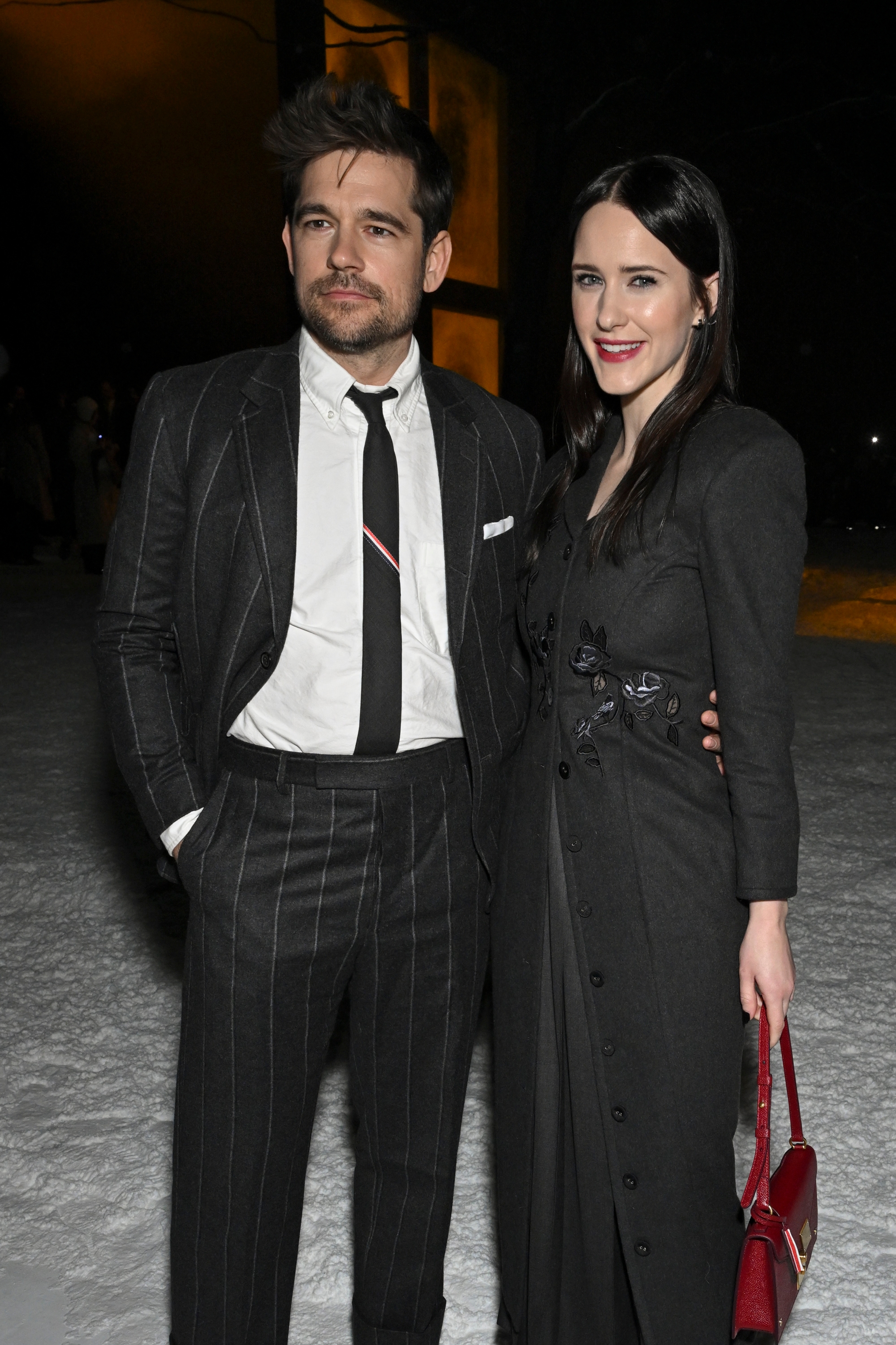 Jason Ralph in a pinstripe suit with a tie and Rachel Brosnahan in a long coat, standing on a snow-covered ground, smiling