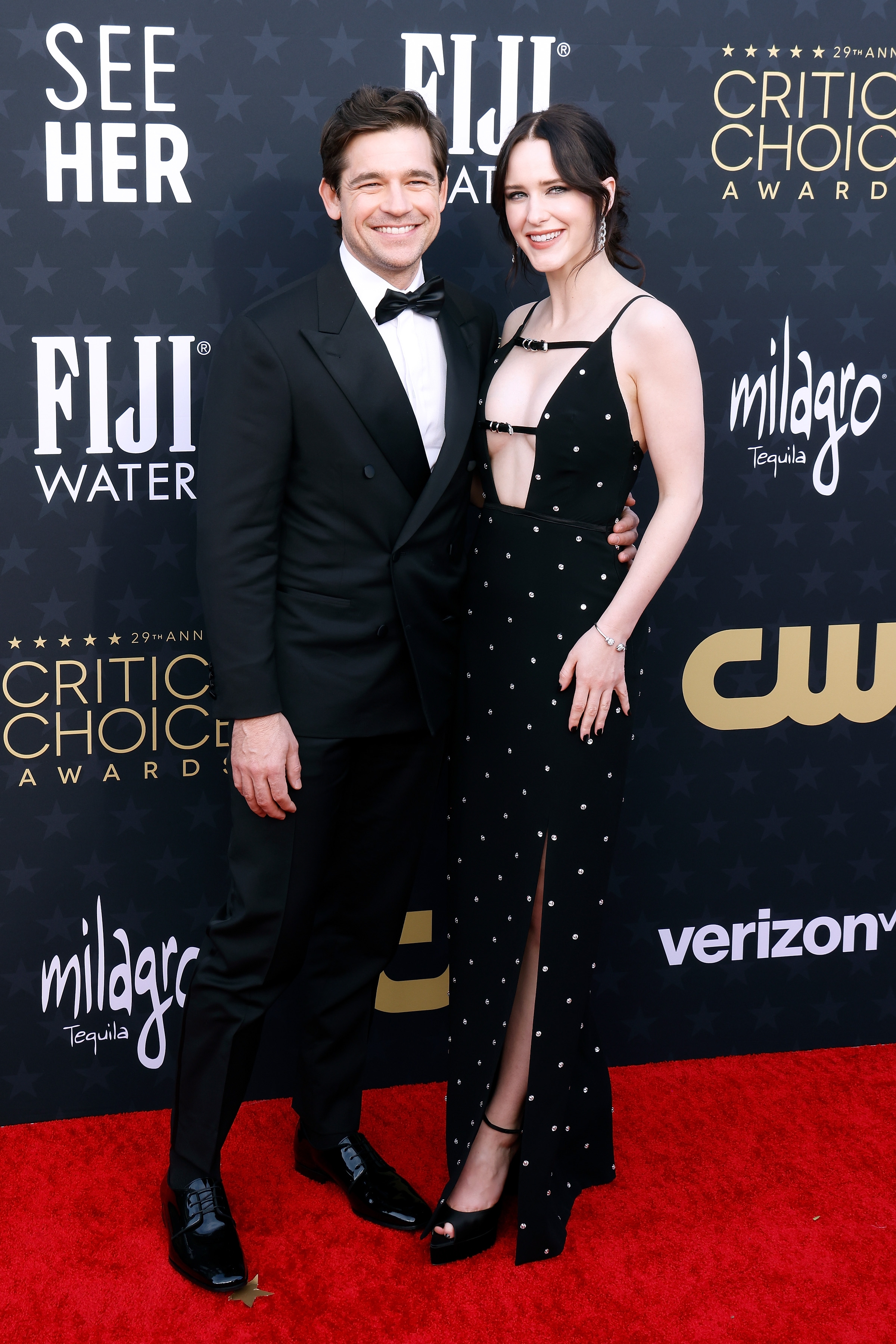 Jason Ralph and Rachel Brosnahan smile as they pose on a red carpet