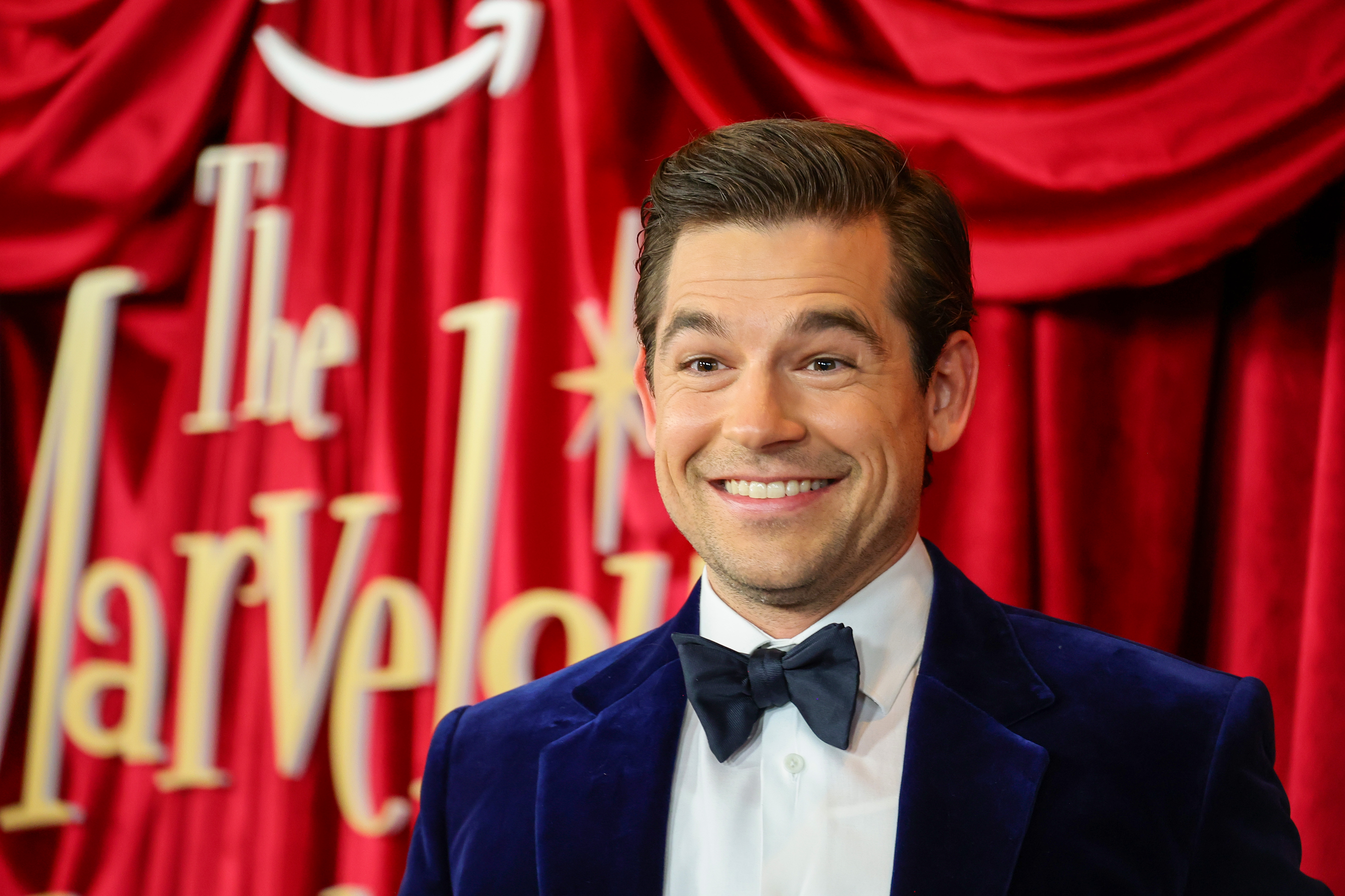 Jason Ralph in a velvet suit with a bow tie smiles on a red carpet backdrop with the word "Marvelous."
