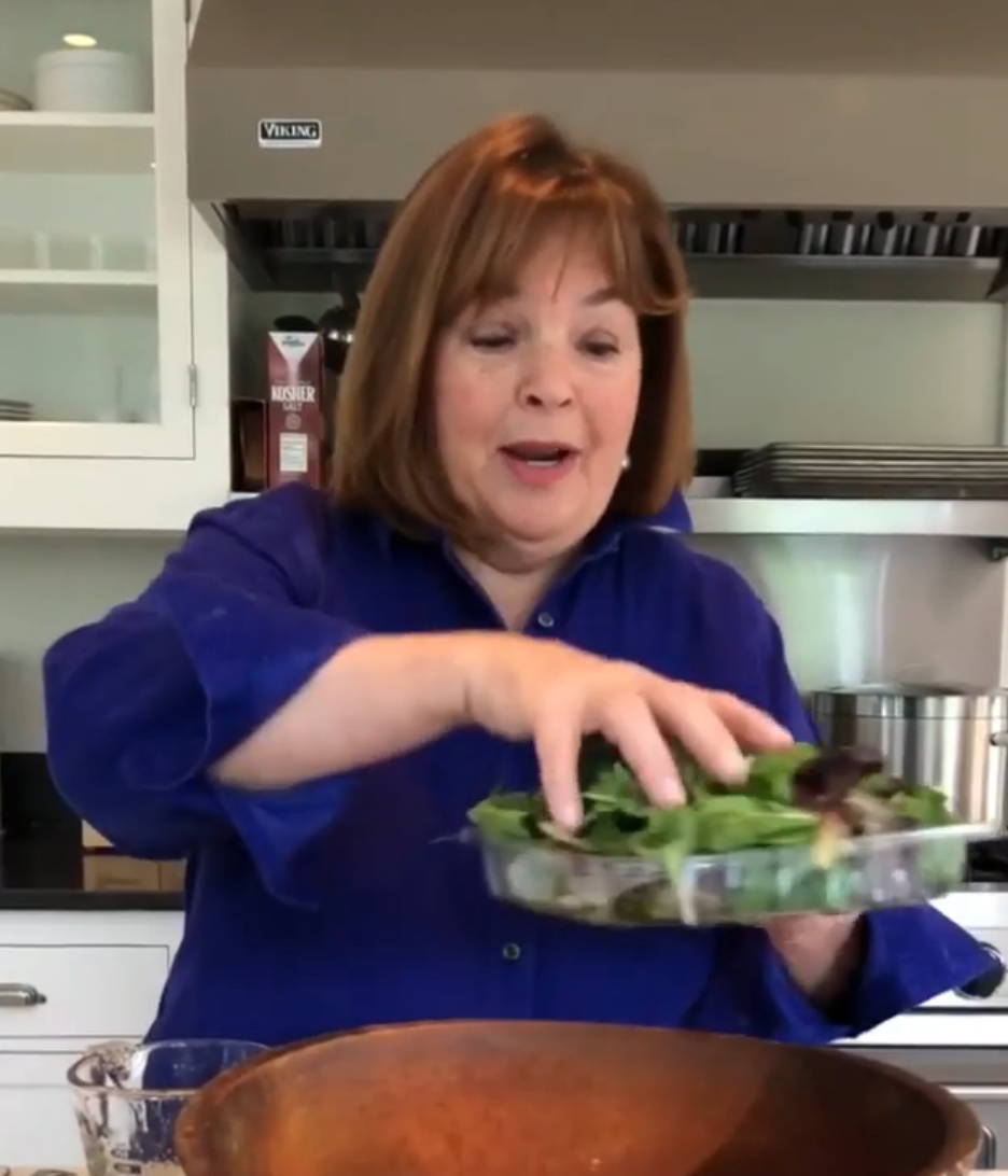 A woman in a kitchen holds a container of mixed greens, preparing to add them to a wooden bowl