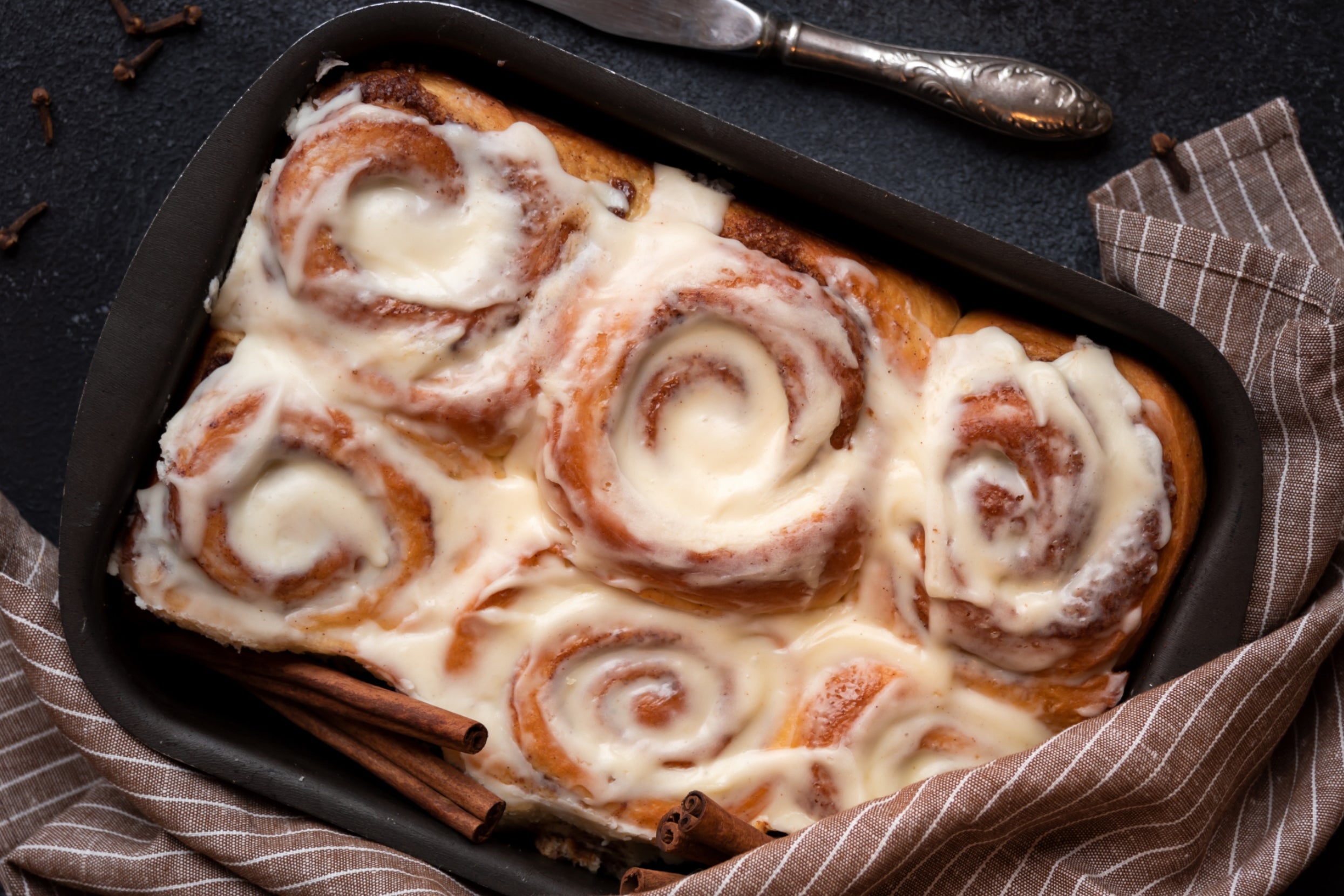 Cinnamon rolls topped with creamy icing in a baking dish, with a towel and cinnamon sticks nearby