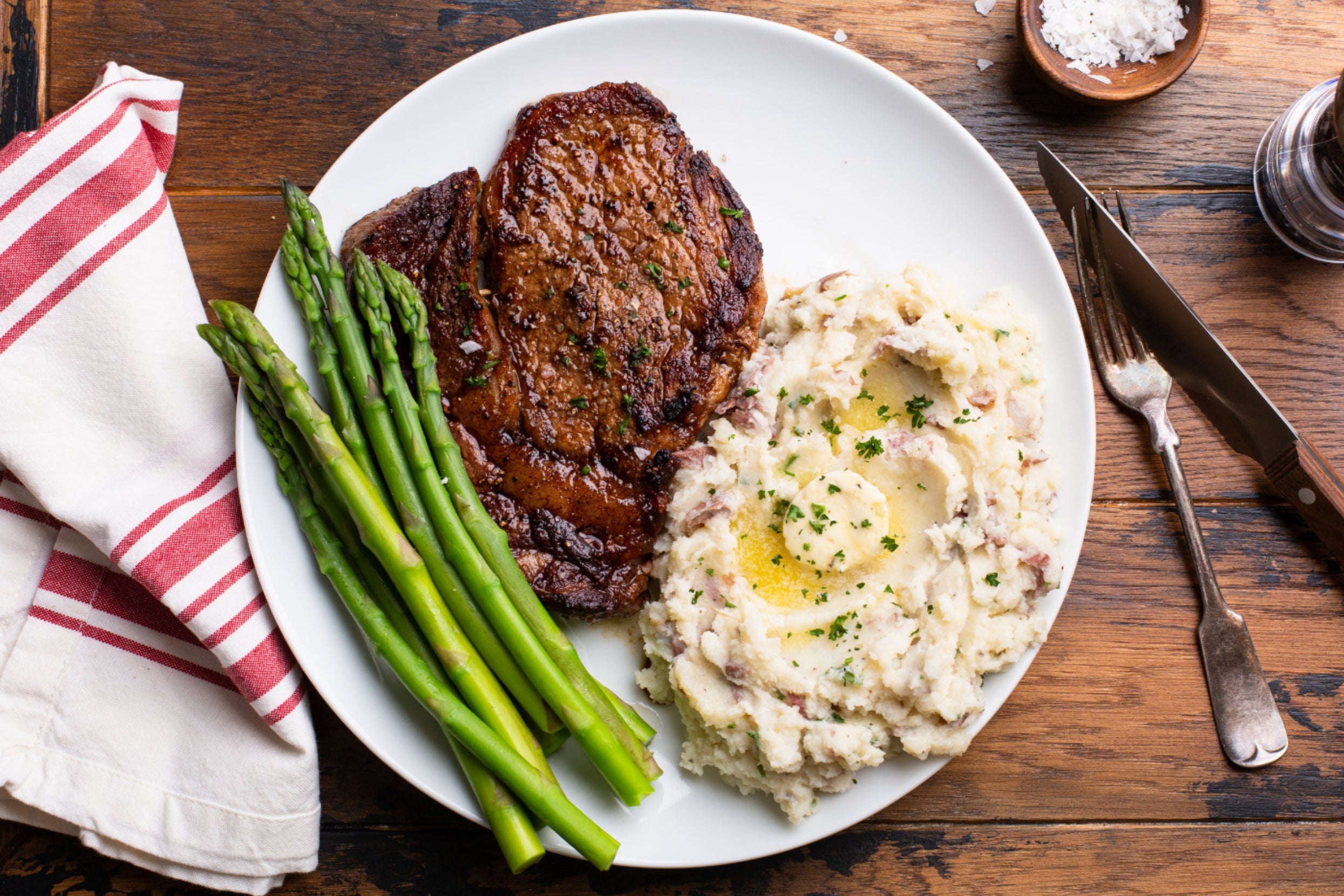 Steak, asparagus, and mashed potatoes with a butter topping on a plate, set on a wooden table with a napkin and cutlery