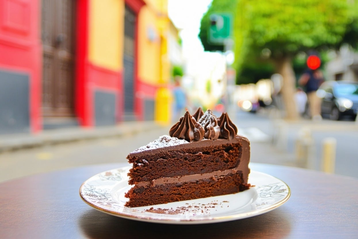A slice of chocolate cake with chocolate frosting and powdered sugar sits on a plate, with a blurred street scene in the background