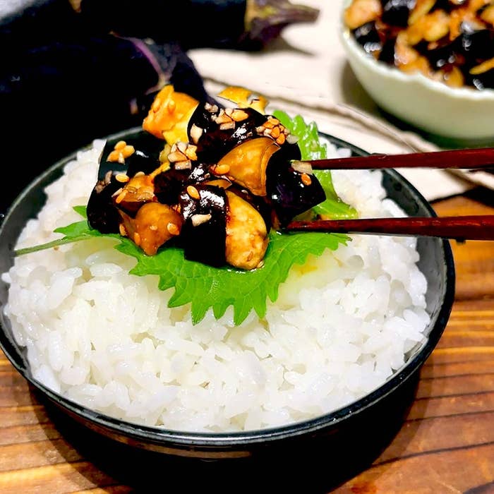 ご飯の上にシソの葉、茄子とごまの料理が盛り付けられ、箸で持ち上げている。
