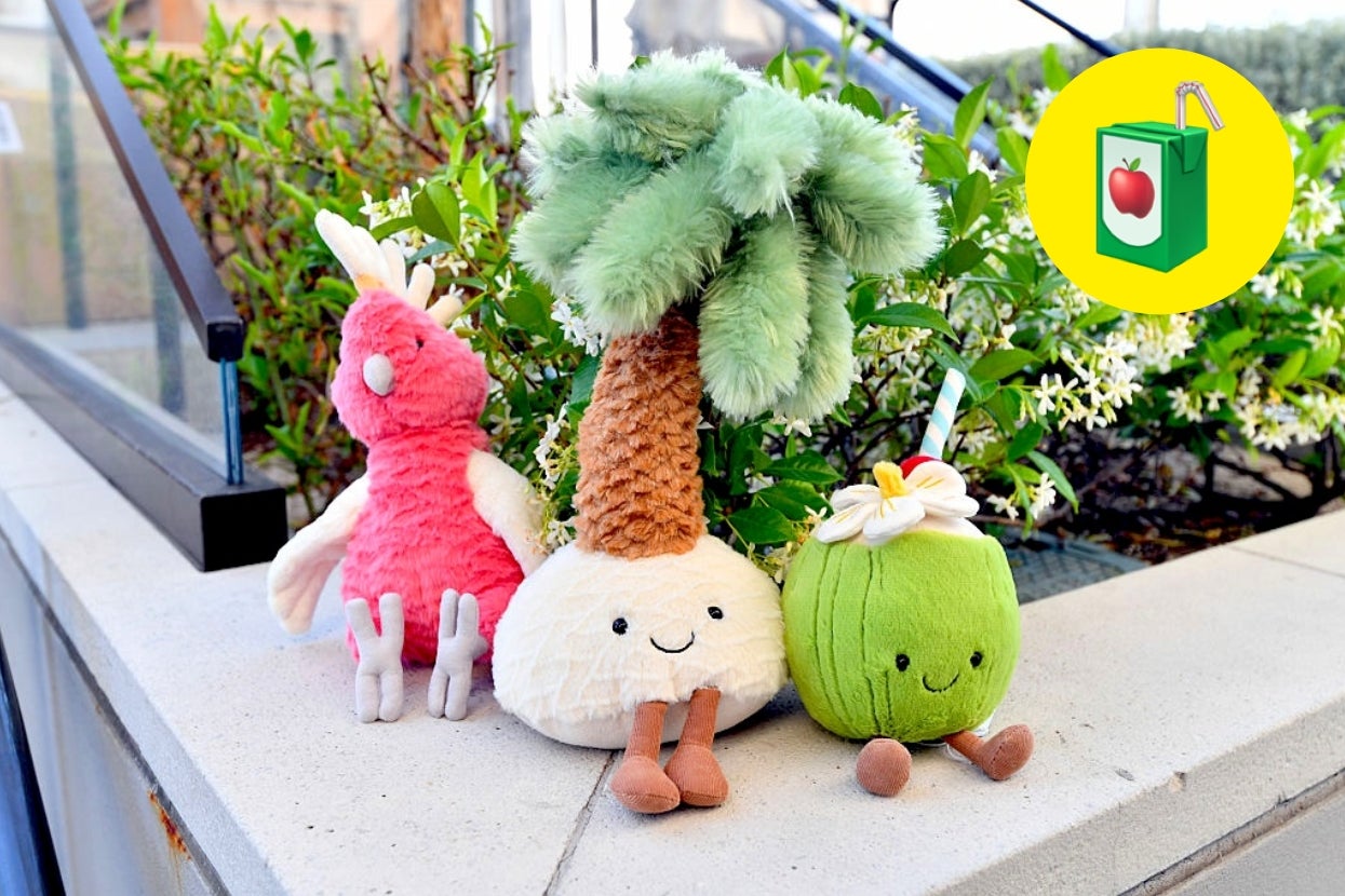Three plush toys: a pink bird, a tree with a smiling face, and a green fruit with a flower, sit on an outdoor ledge near bushes