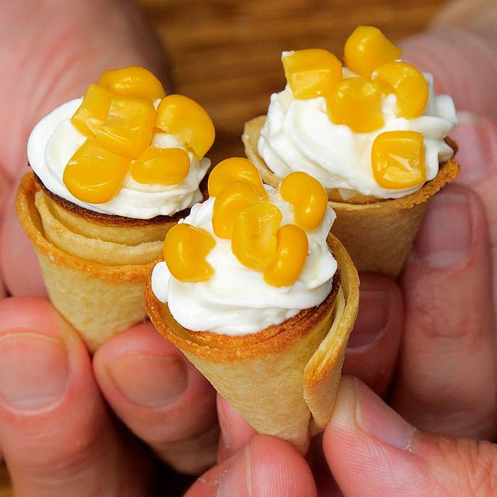 3つの小さな食パンコーンに、ヨーグルトクリームとコーンがトッピングされている。手で持たれている。