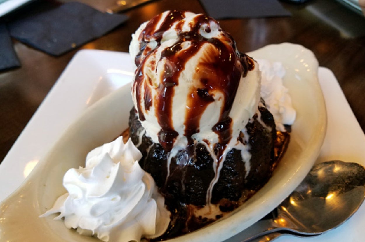 Dessert with ice cream on brownie, drizzled with chocolate sauce, served with dollops of whipped cream on a dish with a spoon