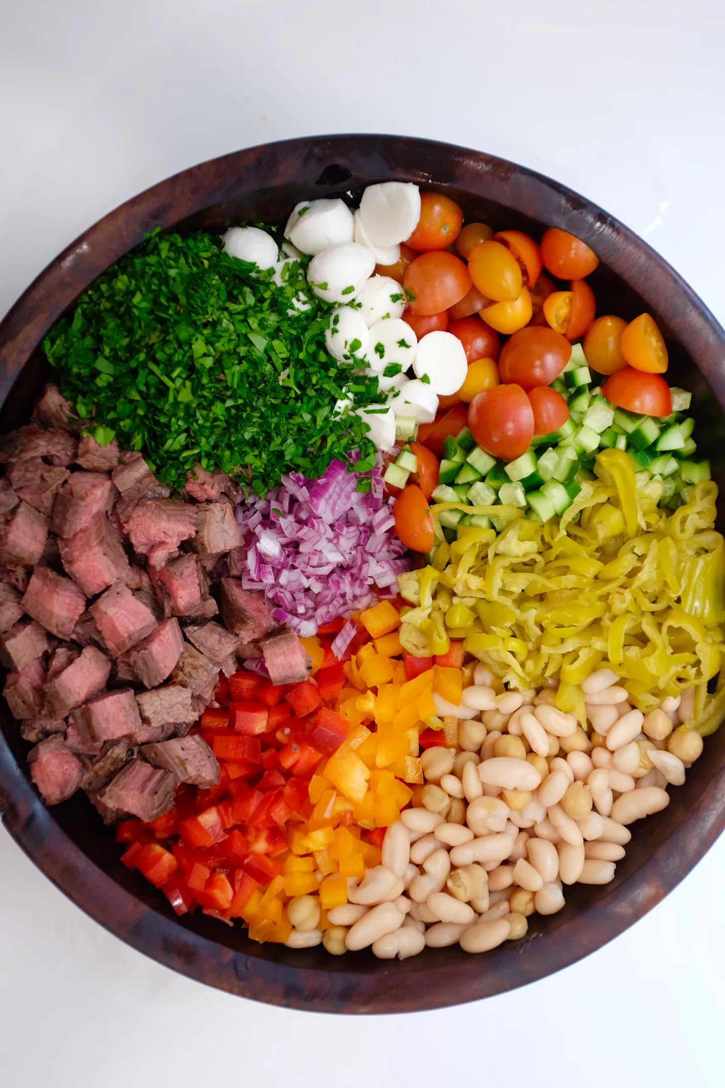 Salad bowl with steak cubes, cherry tomatoes, cucumbers, parsley, mozzarella, onions, bell peppers, pepperoncini, and white beans
