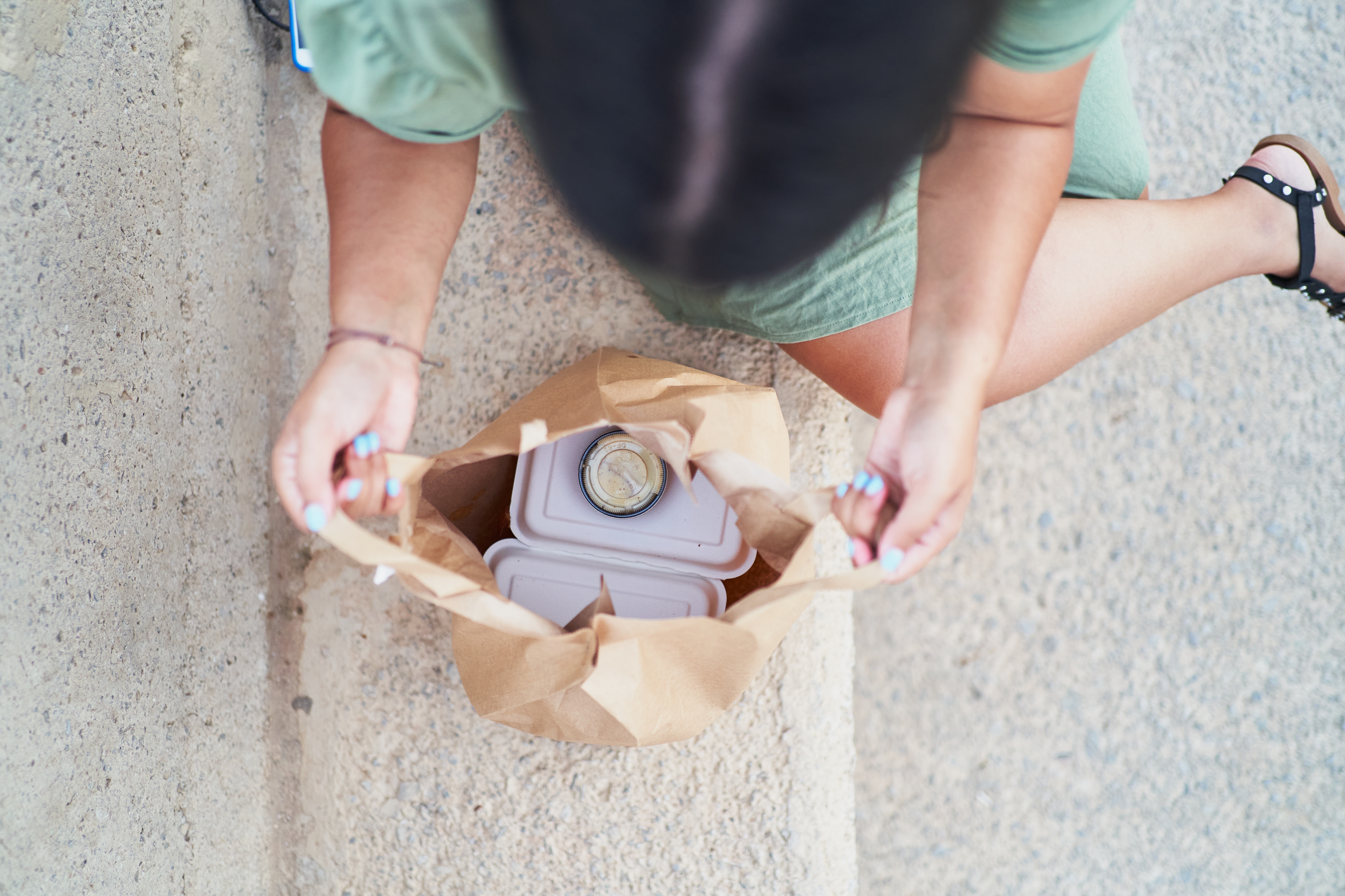 Person sitting on a sidewalk opens a brown paper bag with takeout containers and a drink inside