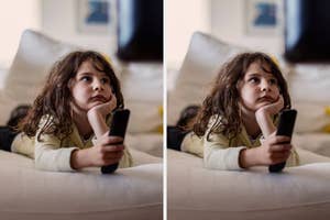 Child lying on a couch holding a remote, gazing at the TV screen, looking thoughtful