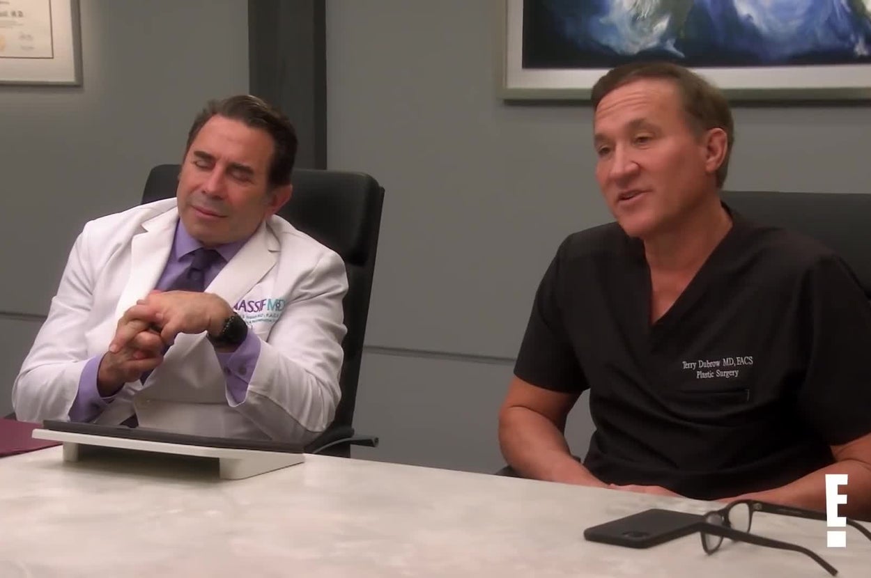 Two doctors sit at a table, one in a white coat and the other in scrubs, engaged in conversation