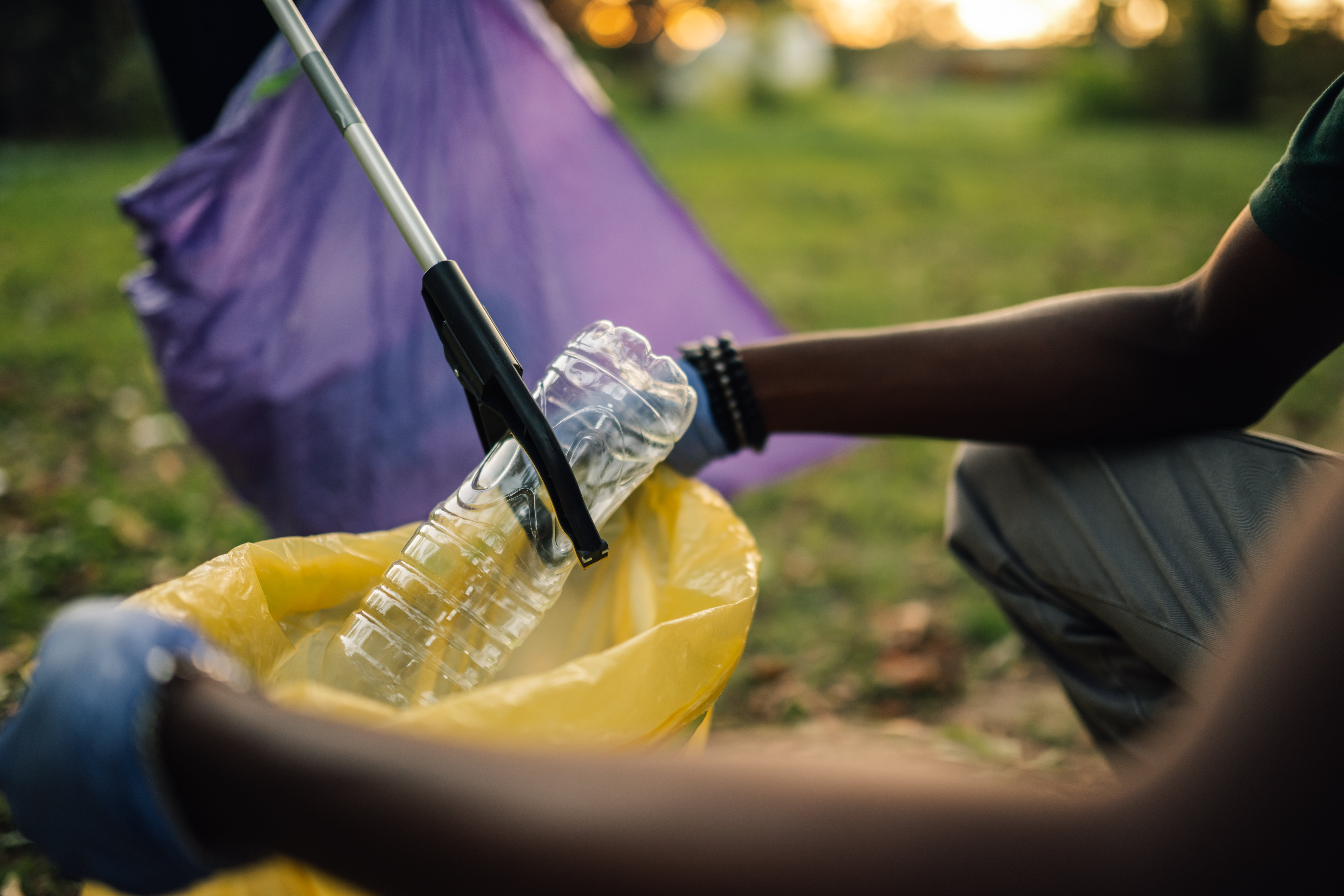 Folk som plukker opp søppel med grabbere, plasserer en plastflaske i en gul pose, og driver med en samfunnsopprydningsinnsats