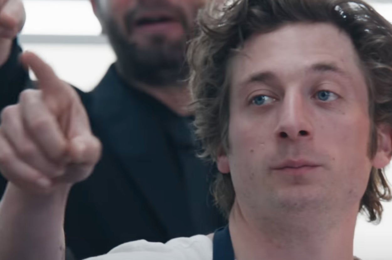 A man with curly hair looks off-camera with a contemplative expression while another person points at something nearby