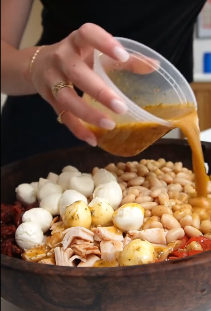 A person pours dressing over a salad with beans, mozzarella, sundried tomatoes, and sliced turkey in a bowl
