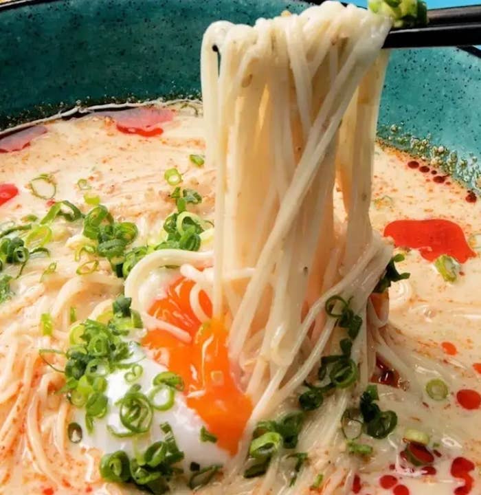 箸で持ち上げられる白い麺、刻みネギ、半熟卵が入ったスープ。